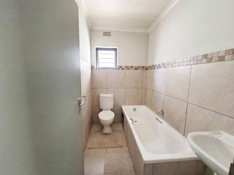 A functional bathroom featuring a bathtub, toilet, and wall-mounted basin with neutral tiled finishes and natural light from a louver window.