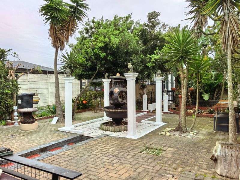 A landscaped garden area featuring a central water feature, decorative columns, and paved pathways surrounded by mature trees and perimeter fencing.