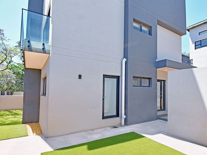 Rear view of a modern townhouse featuring a glass balcony, clean rendered facade, aluminium fixtures, and a low-maintenance garden area with artificial lawn.