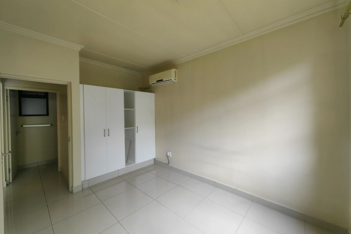 A vacant bedroom featuring tiled flooring, built-in cupboards, and a wall-mounted air conditioning unit, with an adjoining doorway leading to a secondary space.