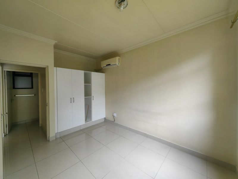 A vacant bedroom featuring tiled flooring, built-in cupboards, and a wall-mounted air conditioning unit, with an adjoining doorway leading to a secondary space.