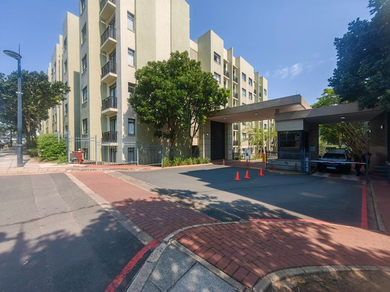 Exterior view of the apartment complex entrance featuring a secure gatehouse, boom gate access, and paved driveway leading to the building facade.
