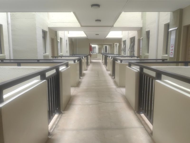 An open-air apartment complex hallway featuring concrete flooring and metal safety railings leading to multiple unit entrances.