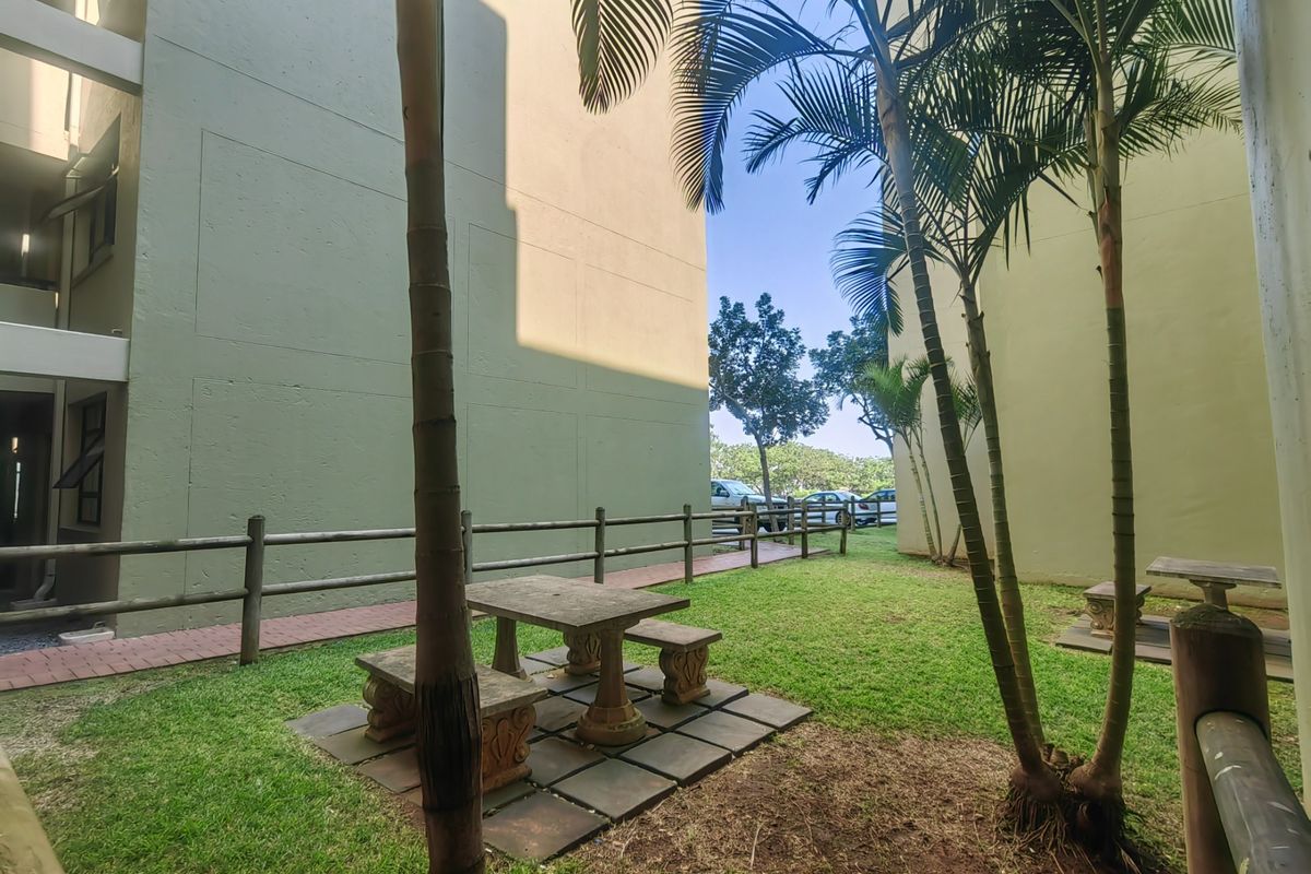 A communal garden area between apartment blocks featuring a paved walkway, timber fencing, and a concrete outdoor seating area.