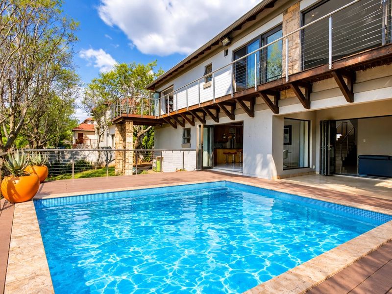 A well-maintained outdoor pool area featuring timber decking, stone accents, and a balcony overlooking the swimming pool with modern stainless steel balustrades.
