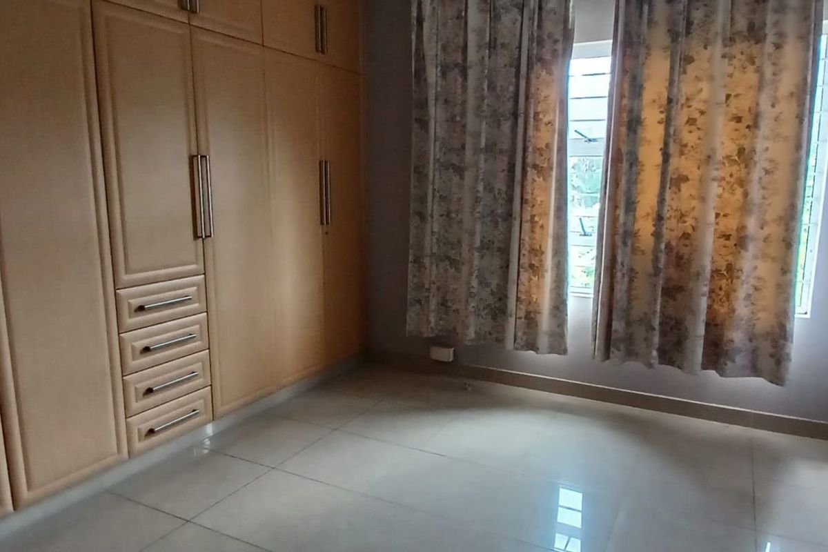 A clean, empty bedroom featuring extensive built-in wooden cupboards, neutral tiled flooring, and a window fitted with security bars.