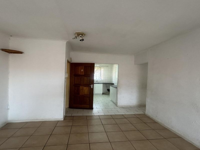 An empty open-plan living area with tiled flooring, leading through a wooden door into a kitchen space.
