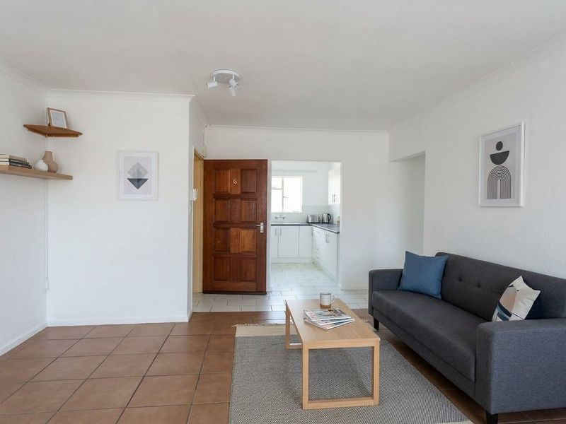 A bright living room featuring tiled flooring and a direct view into the adjacent kitchen area through a wooden entrance door.