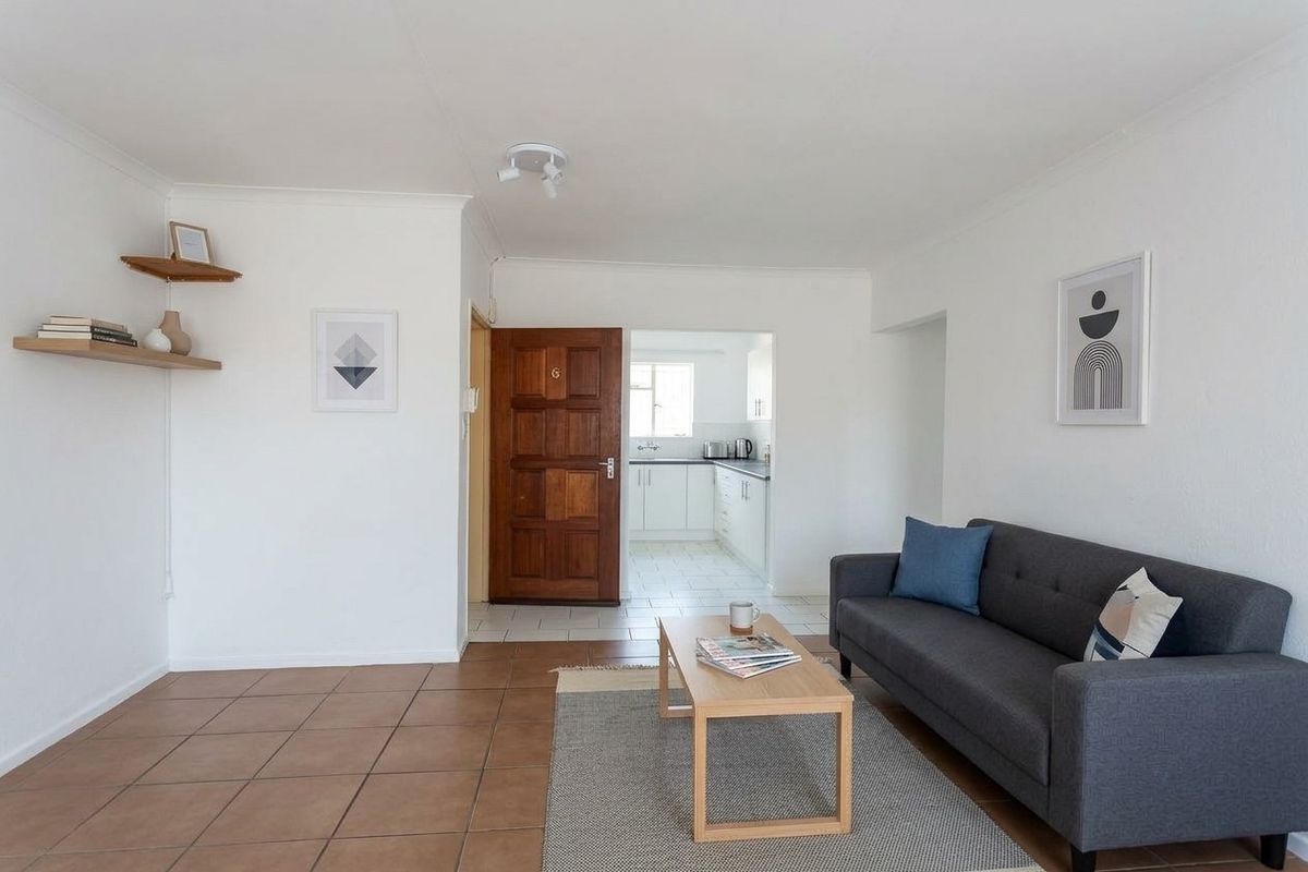 A bright living room featuring tiled flooring and a direct view into the adjacent kitchen area through a wooden entrance door.