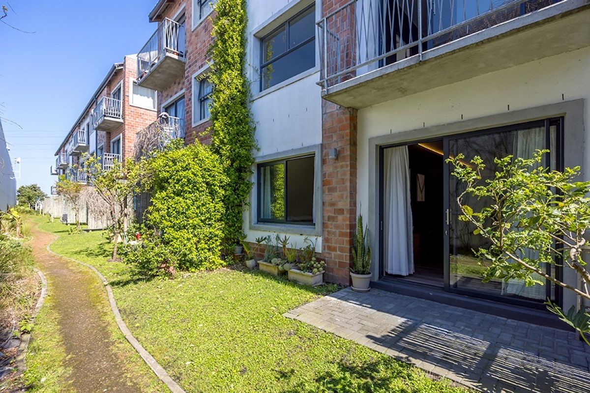 Rear view of the apartment complex featuring brick facade, private ground floor patio with sliding door access, and a landscaped garden walkway.
