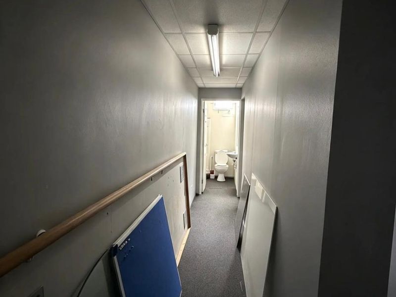 A narrow commercial hallway with carpet flooring, suspended ceiling, and fluorescent lighting, leading towards a small restroom area.