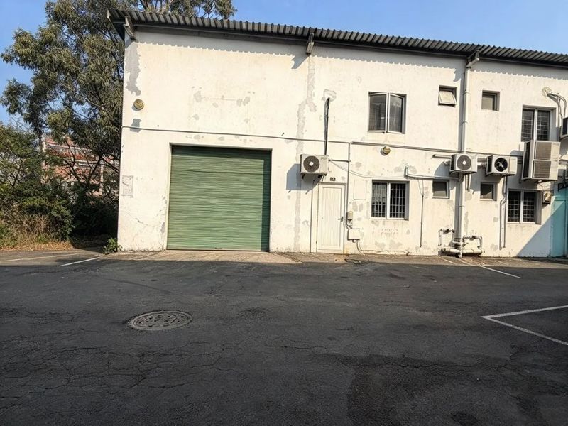 Rear exterior view of a commercial building featuring a roller shutter door, asphalt parking area, and multiple external air conditioning units.