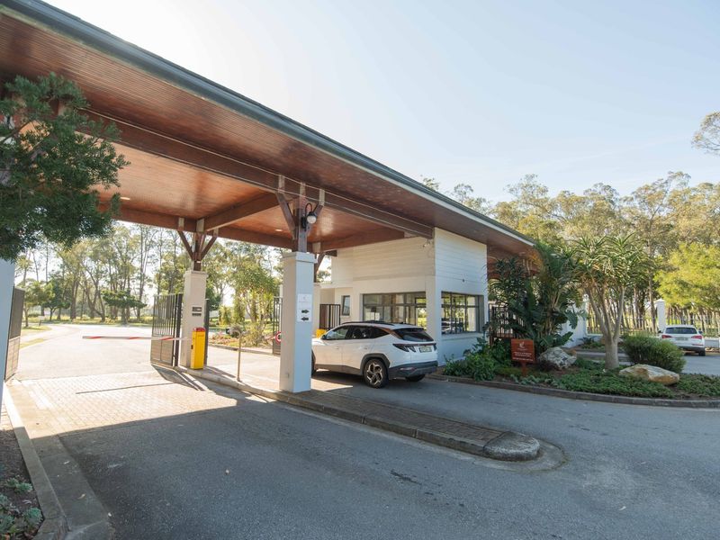 A well-maintained security gatehouse entrance featuring a large timber-clad canopy, boom gate access, and landscaped garden beds.