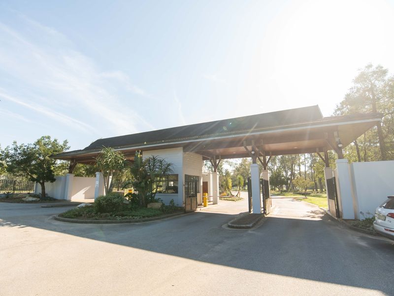 A well-maintained security gatehouse entrance featuring a paved driveway, perimeter walls, and a covered timber roof structure at the estate access point.