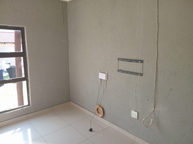 An empty bedroom featuring tiled flooring, an aluminium window, and wall-mounted infrastructure including a TV bracket and exposed electrical wiring.