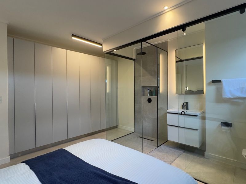 Modern bedroom featuring extensive built-in cupboards and a sleek, glass-enclosed en-suite bathroom with a walk-in shower and contemporary vanity.
