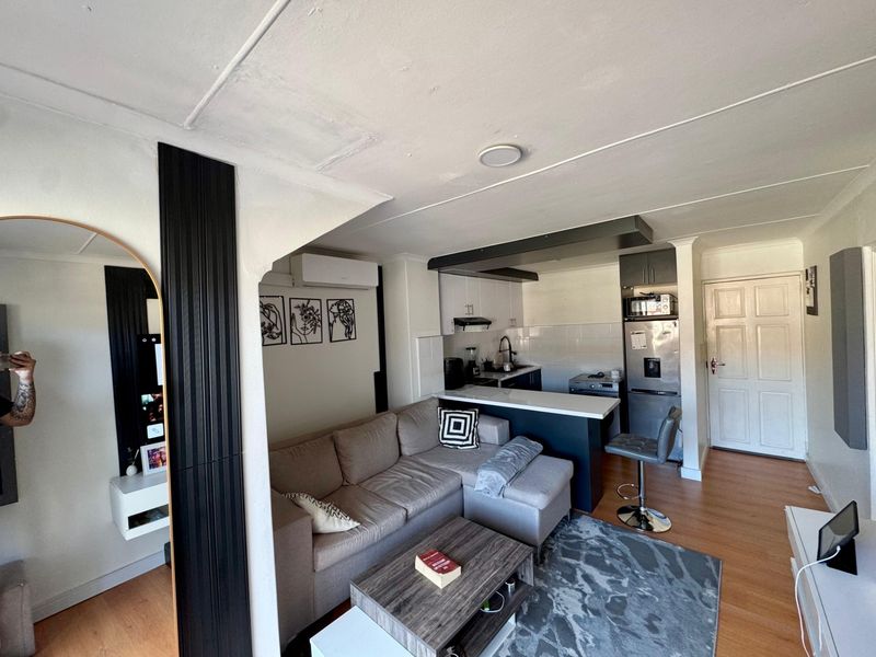 Open-plan living area featuring a compact kitchen with breakfast bar, laminate flooring, and an integrated air conditioning unit.