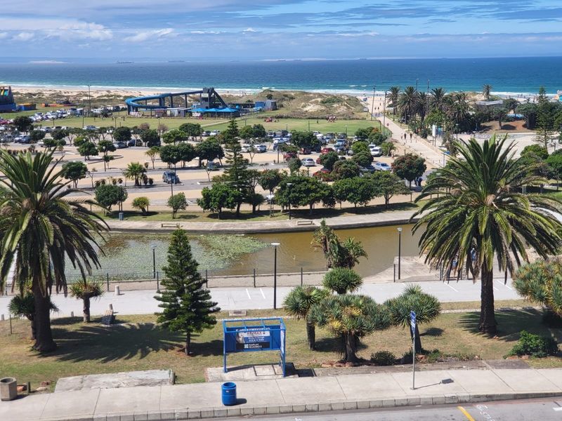 Elevated coastal view overlooking a public park, parking area, and the ocean beach in the distance.
