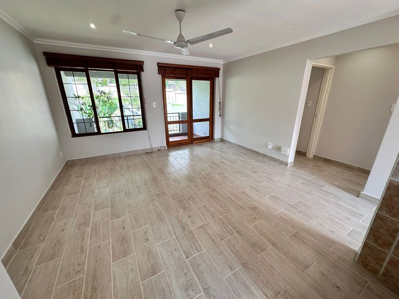 A bright, empty living room featuring modern wood-look tiled flooring, recessed ceiling lights, a ceiling fan, and access to an outdoor area.