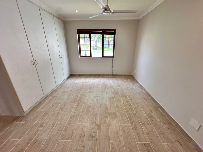 A bright, empty bedroom featuring modern wood-look floor tiles, built-in cupboards, and a ceiling fan, offering a clean and neutral space.