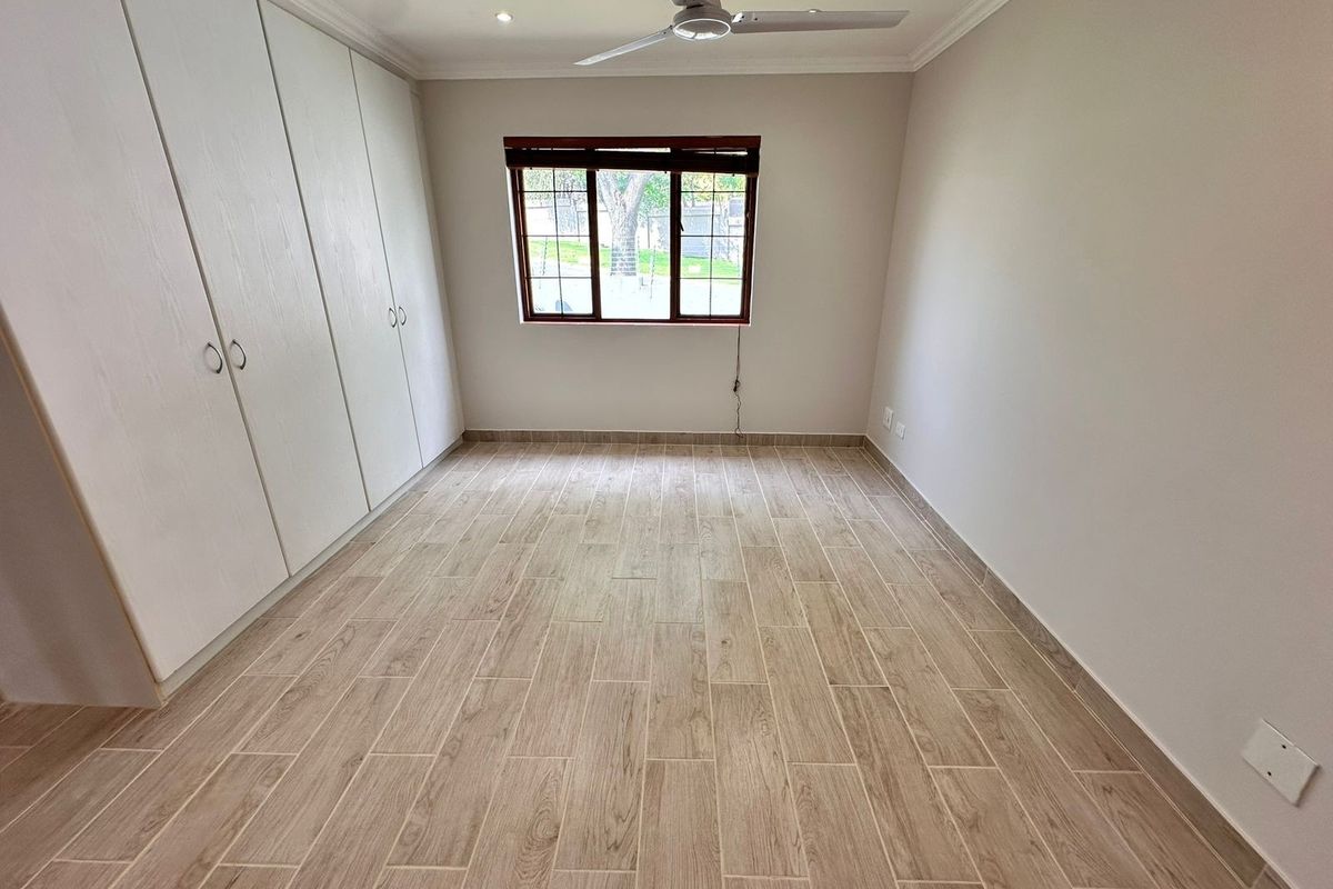 A bright, empty bedroom featuring modern wood-look floor tiles, built-in cupboards, and a ceiling fan, offering a clean and neutral space.
