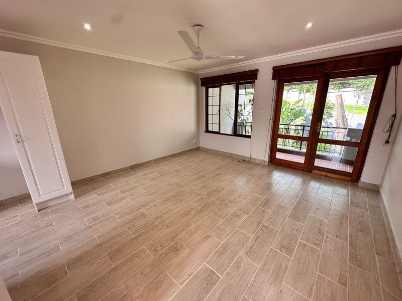 A bright, empty living room featuring wood-look tiled flooring, a ceiling fan, built-in storage, and direct access to a balcony through wooden framed glass doors.