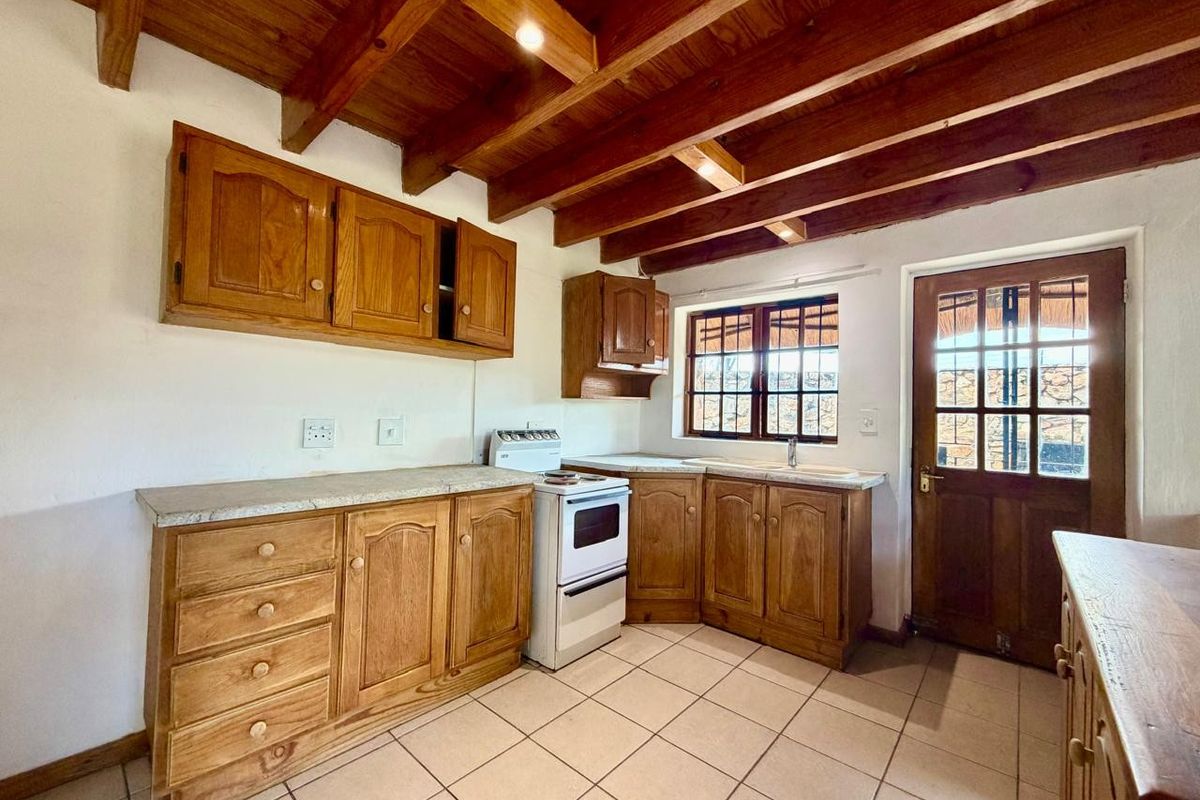A rustic kitchen featuring timber cabinetry, exposed ceiling beams, tiled flooring, and a freestanding stove with direct access to the exterior.