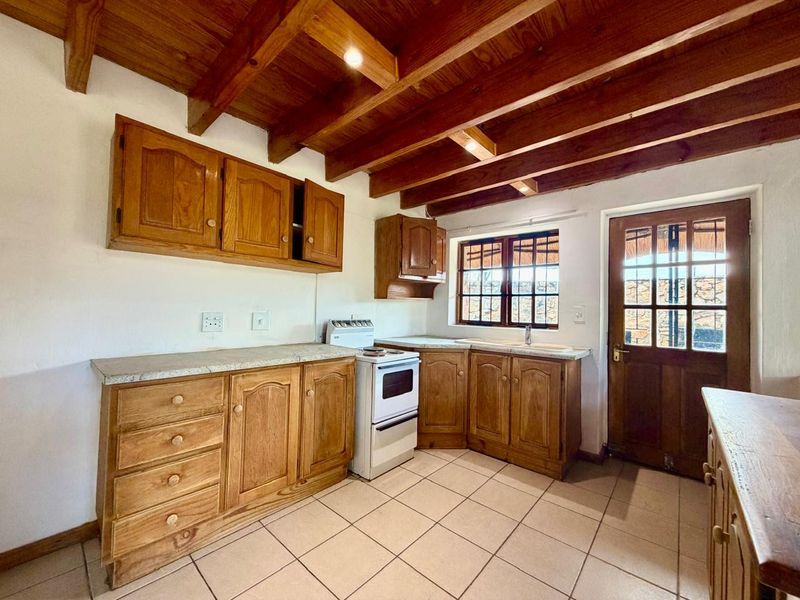A rustic kitchen featuring timber cabinetry, exposed ceiling beams, tiled flooring, and a freestanding stove with direct access to the exterior.
