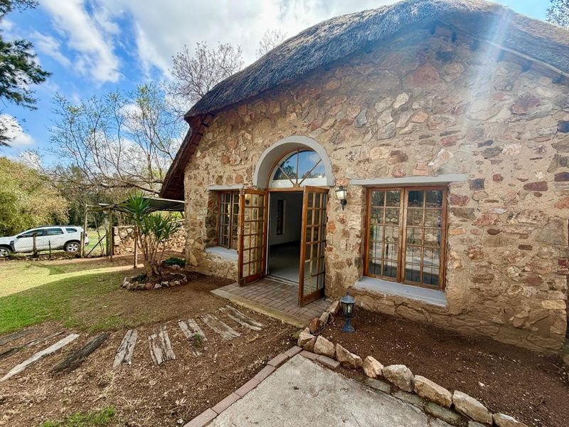 Charming cottage exterior featuring traditional stone masonry walls, a thatched roof, and arched timber-framed entrance doors opening onto a garden setting.