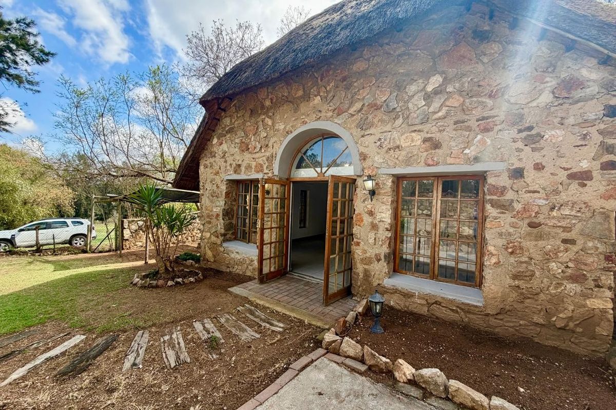 Charming cottage exterior featuring traditional stone masonry walls, a thatched roof, and arched timber-framed entrance doors opening onto a garden setting.