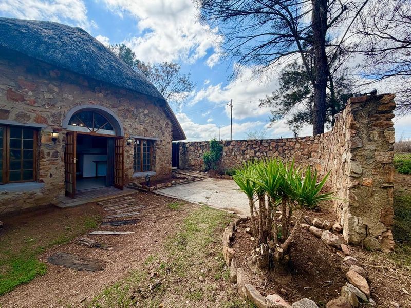 Rear view of a charming stone-clad cottage featuring a traditional thatched roof, a private paved patio, and a rustic stone boundary wall.