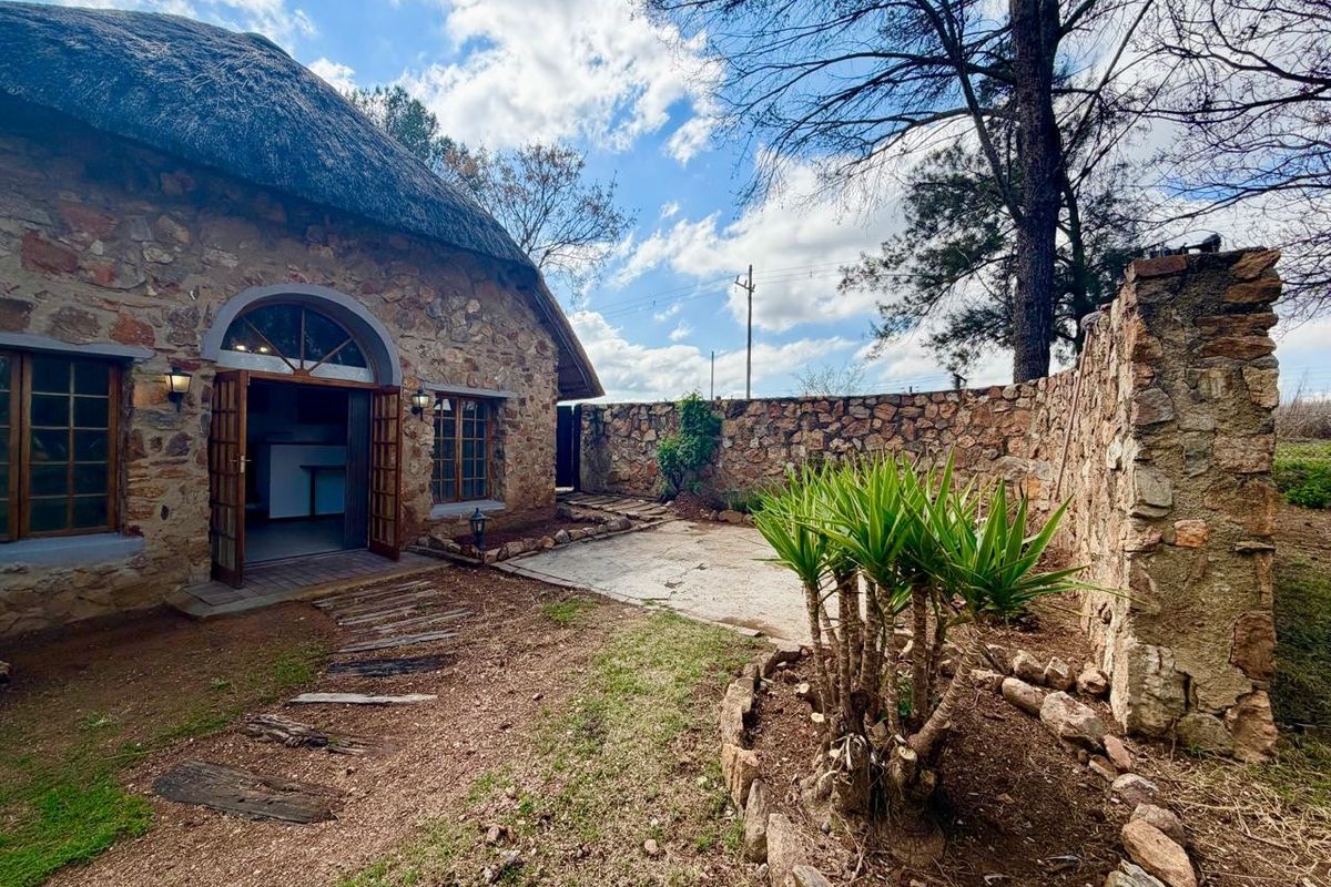 Rear view of a charming stone-clad cottage featuring a traditional thatched roof, a private paved patio, and a rustic stone boundary wall.
