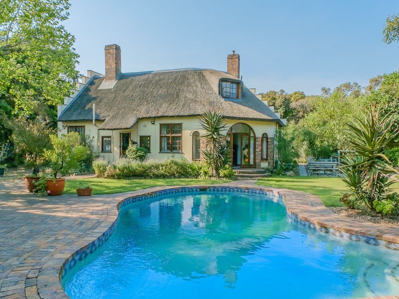 A charming rear exterior view featuring a classic thatched roof house, a well-maintained swimming pool, and a lush, landscaped garden setting.