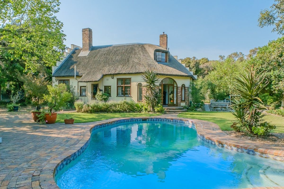 A charming rear exterior view featuring a classic thatched roof house, a well-maintained swimming pool, and a lush, landscaped garden setting.