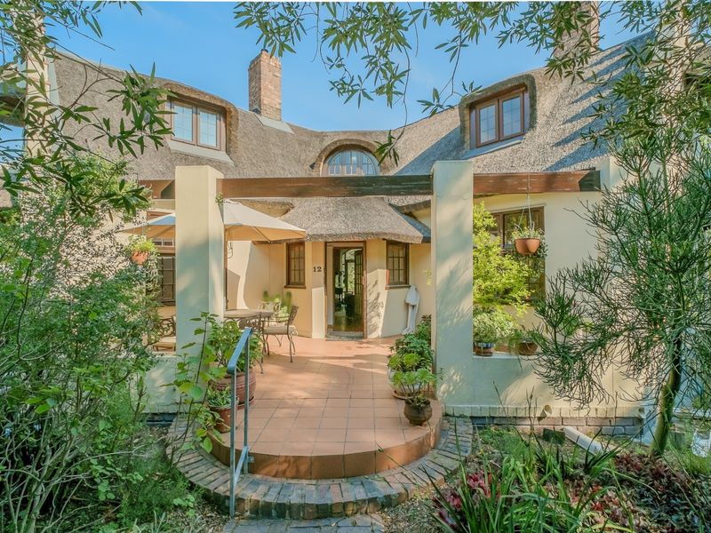 A charming rear exterior view featuring a traditional thatched roof, a covered tiled patio with a timber pergola, and established garden surroundings.