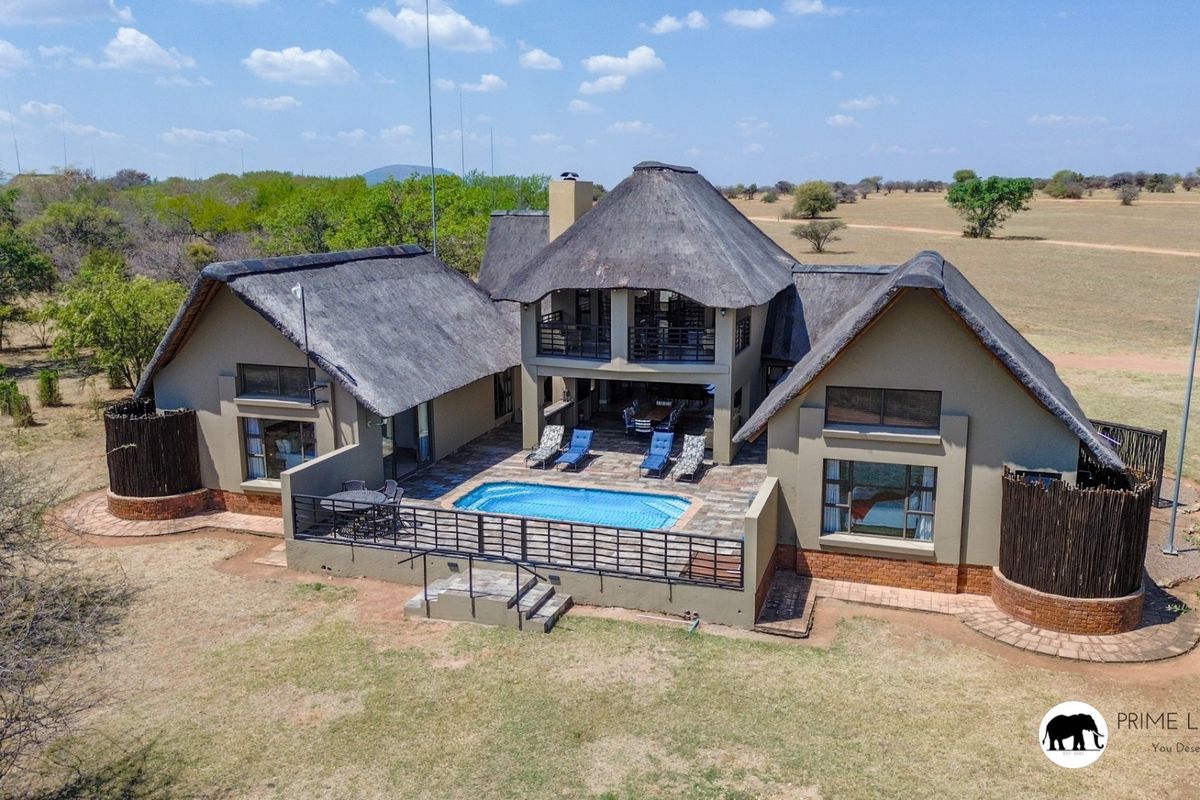 Aerial view of a large thatched-roof house featuring a swimming pool, paved patio area, and balcony, set within a spacious natural landscape.