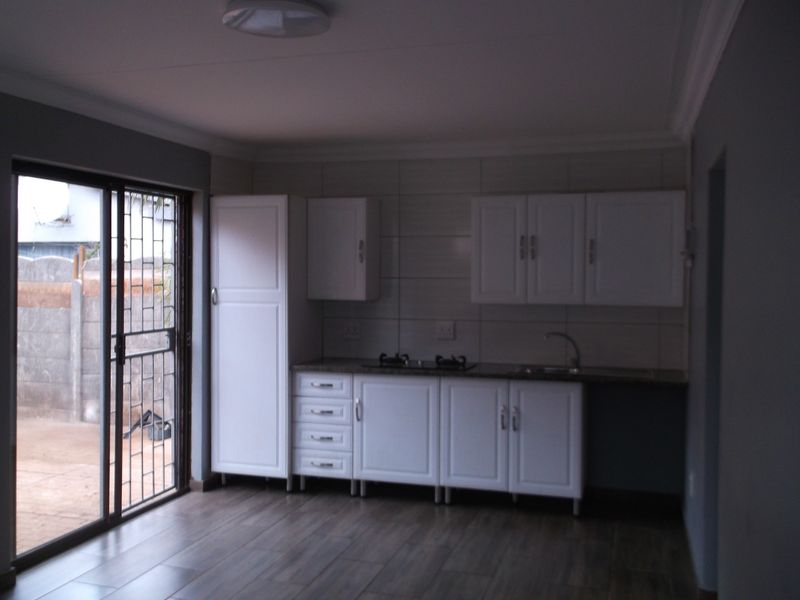 A compact, modern kitchen featuring white cabinetry, a built-in hob, sink, and laminate flooring, with direct access to the exterior via a sliding glass door.