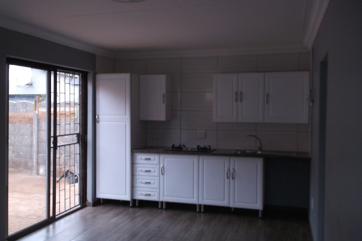 A compact, modern kitchen featuring white cabinetry, a built-in hob, sink, and laminate flooring, with direct access to the exterior via a sliding glass door.