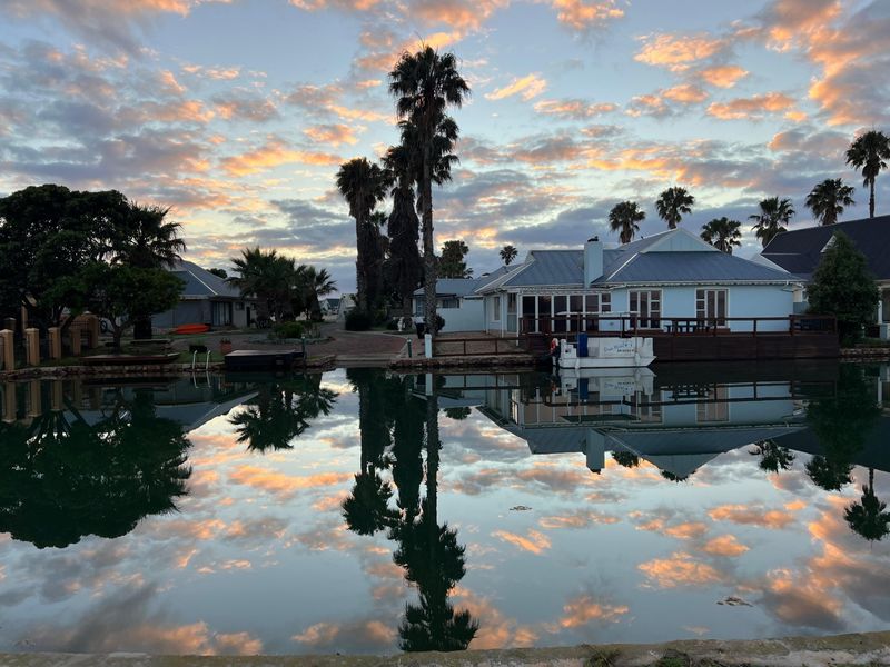 A scenic rear view of a waterfront residential property featuring a timber deck, metal roofing, and established palm trees at sunset.