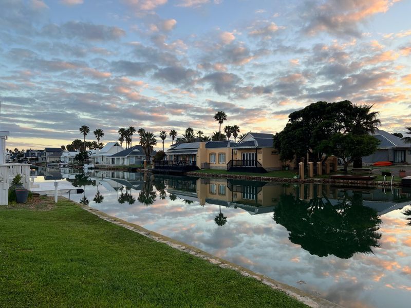 A scenic view of a tranquil canal lined with residential properties, mature trees, and well-maintained lawns under a beautiful sunset sky.