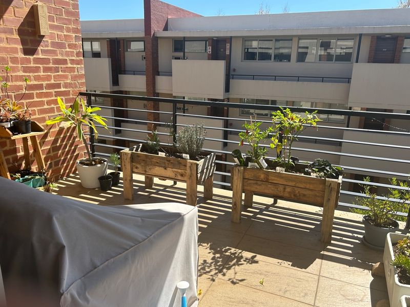 A sunny apartment balcony featuring tiled flooring, a metal balustrade, and an exposed brick wall, overlooking the complex courtyard.