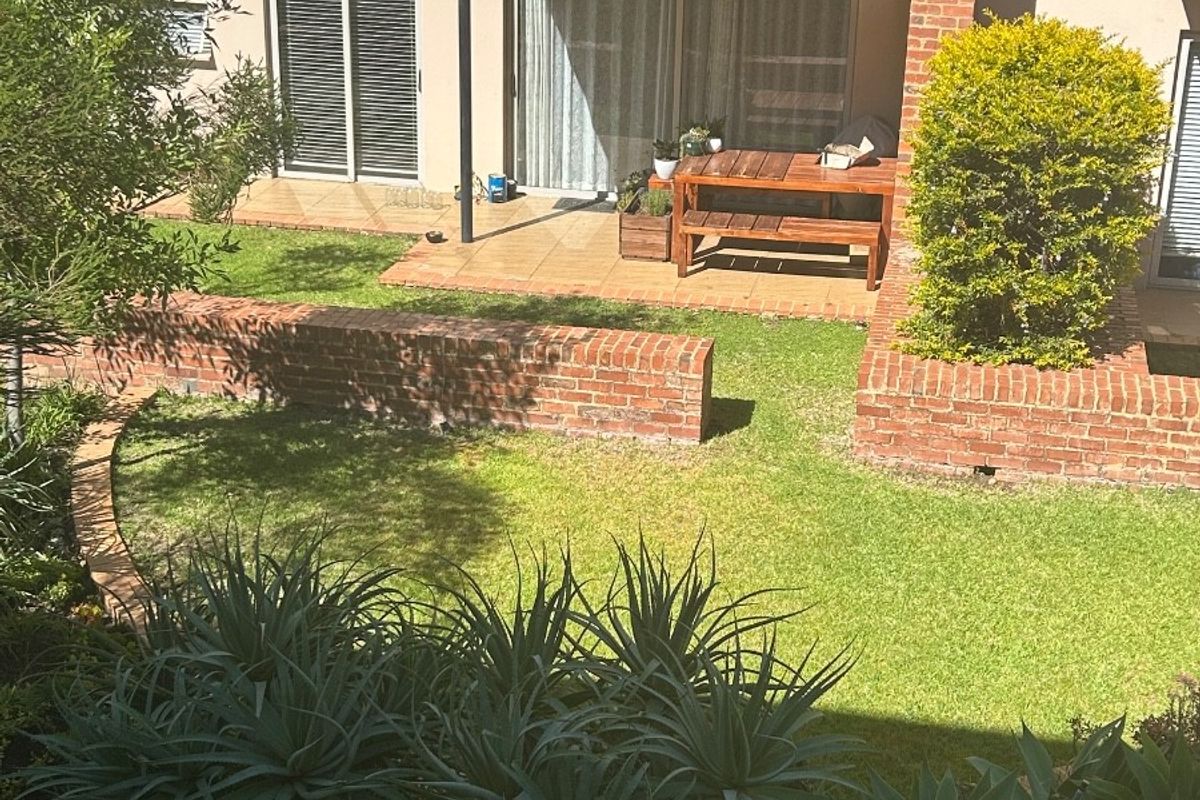 A rear view of the apartment featuring a private garden, brick retaining walls, a tiled patio area, and an upper-level balcony with metal railings.