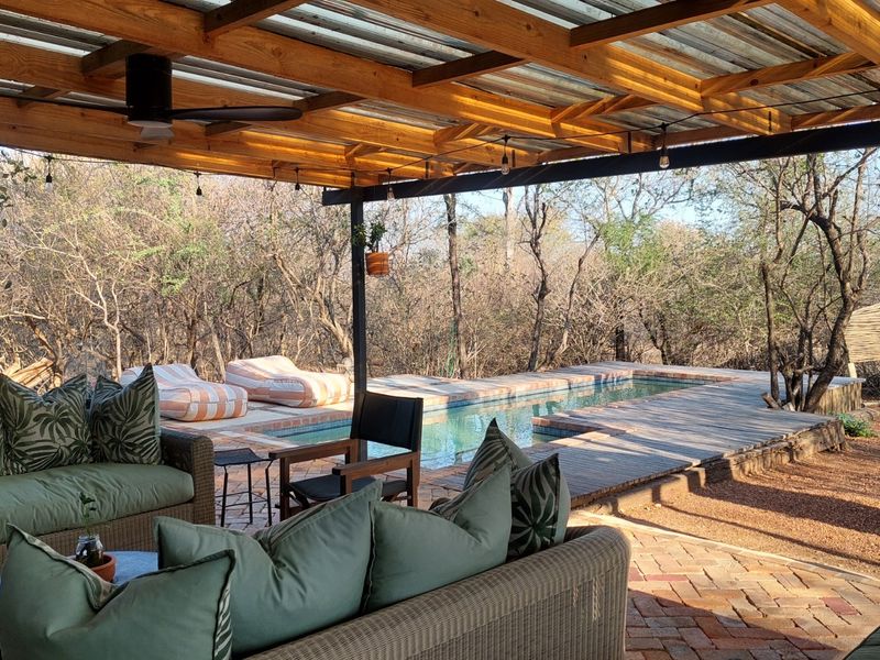 A spacious covered patio featuring a timber pergola and ceiling fan, overlooking a swimming pool with timber decking and natural bushveld surroundings.