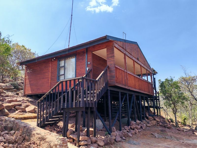 A raised timber cabin featuring a wrap-around deck, corrugated metal roofing, and elevated stilt foundations set on a rocky hillside.