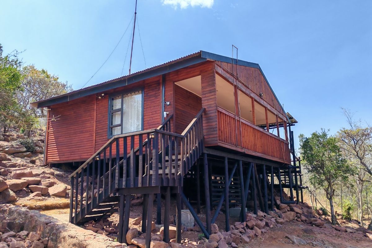 A raised timber cabin featuring a wrap-around deck, corrugated metal roofing, and elevated stilt foundations set on a rocky hillside.
