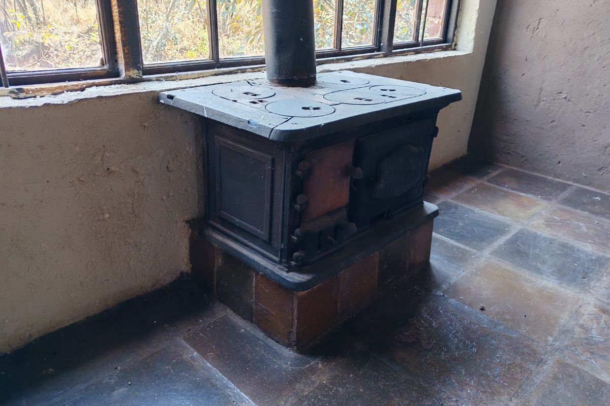 A rustic living area featuring a vintage cast iron wood-burning stove, tiled flooring, and large steel-framed windows overlooking the surrounding landscape.