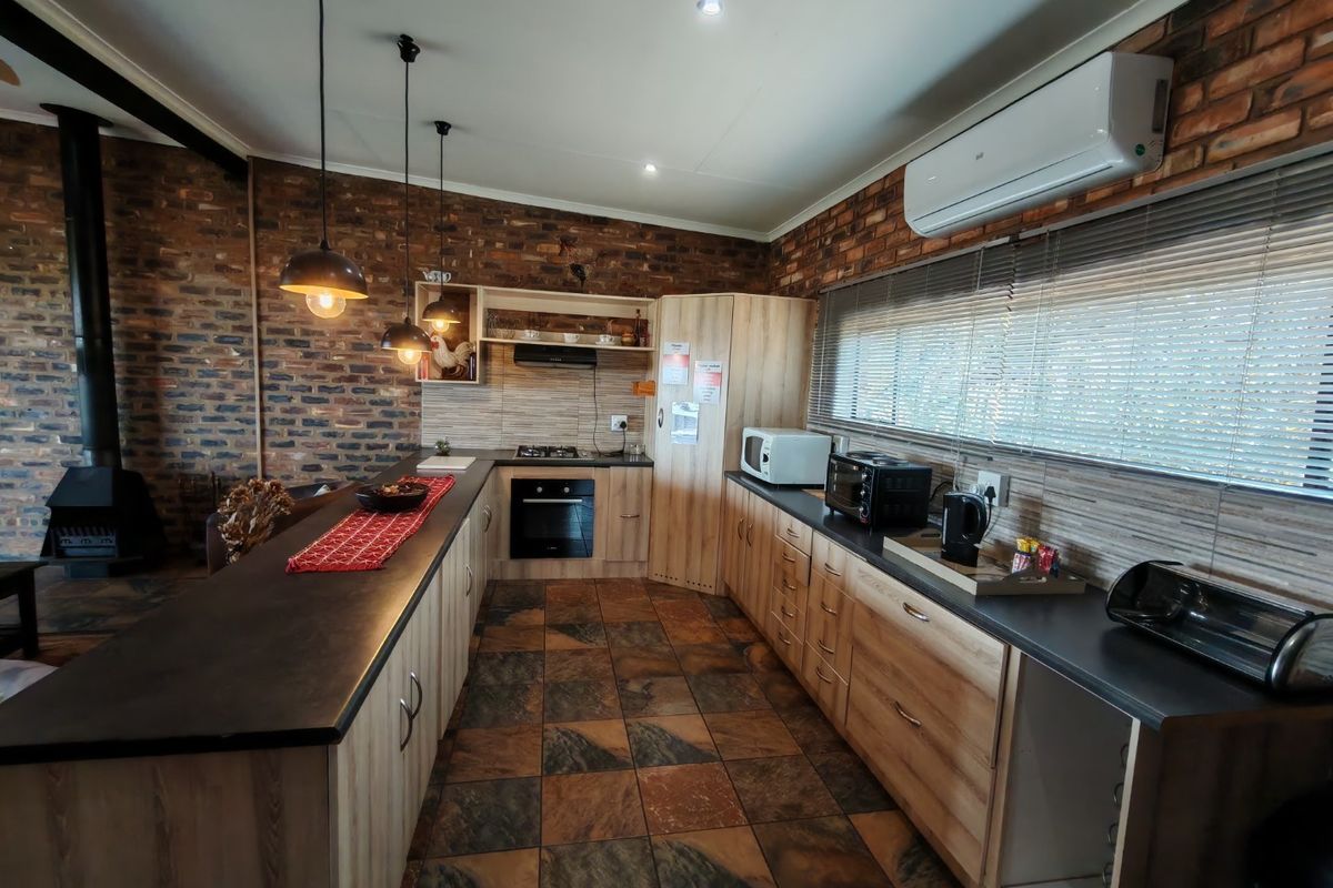A well-appointed kitchen featuring wood-look cabinetry, dark countertops, exposed brick walls, and a cozy wood-burning fireplace, complemented by modern lighting and air conditioning.