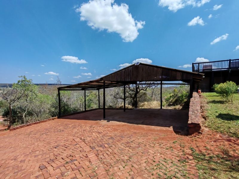 An open-sided covered patio area featuring brick paving and a stone retaining wall, set against a natural landscape backdrop.
