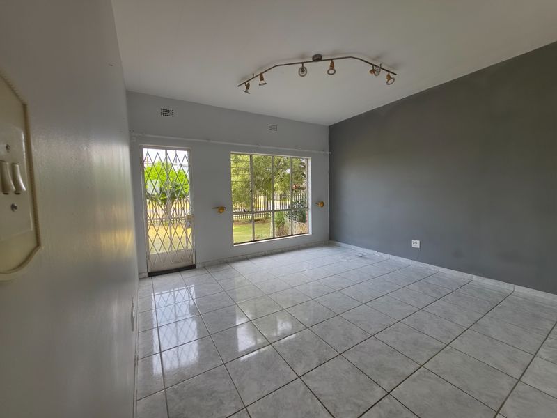 A bright, empty living room featuring neutral tiled flooring, a security gate, and a large window providing natural light and garden views.