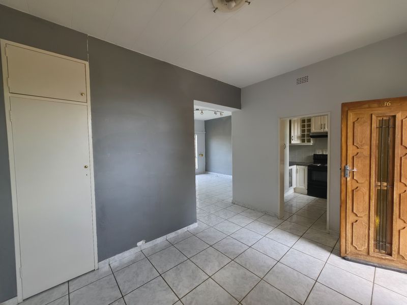 An empty living area featuring tiled flooring, a built-in cupboard, and a wooden entrance door, with a view through to the kitchen.